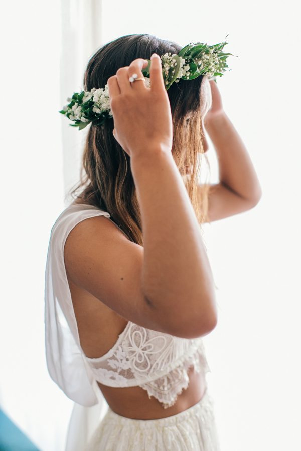 Beautiful boho bride putting on handmade green flower wreath and checking her reflection in the mirror during bridal preparations for a beach ceremony.