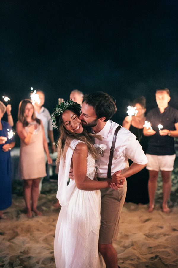 Professional photo of bride and groom during their first dance in the sand surrounded by happy wedding guests holding sparklers. The couple is dancing and laughing.