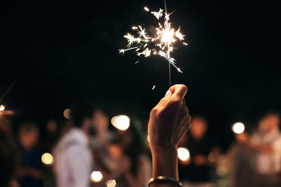 Closeup image of a guest's hand stretched out in the air and holding a sparkler with the wedding couple having their first dance in the background during the wedding reception.