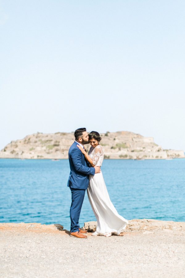 Professional wedding photosession in Elounda Crete, bride and groom walking and posing with Spinalonga island and sea view in the background.