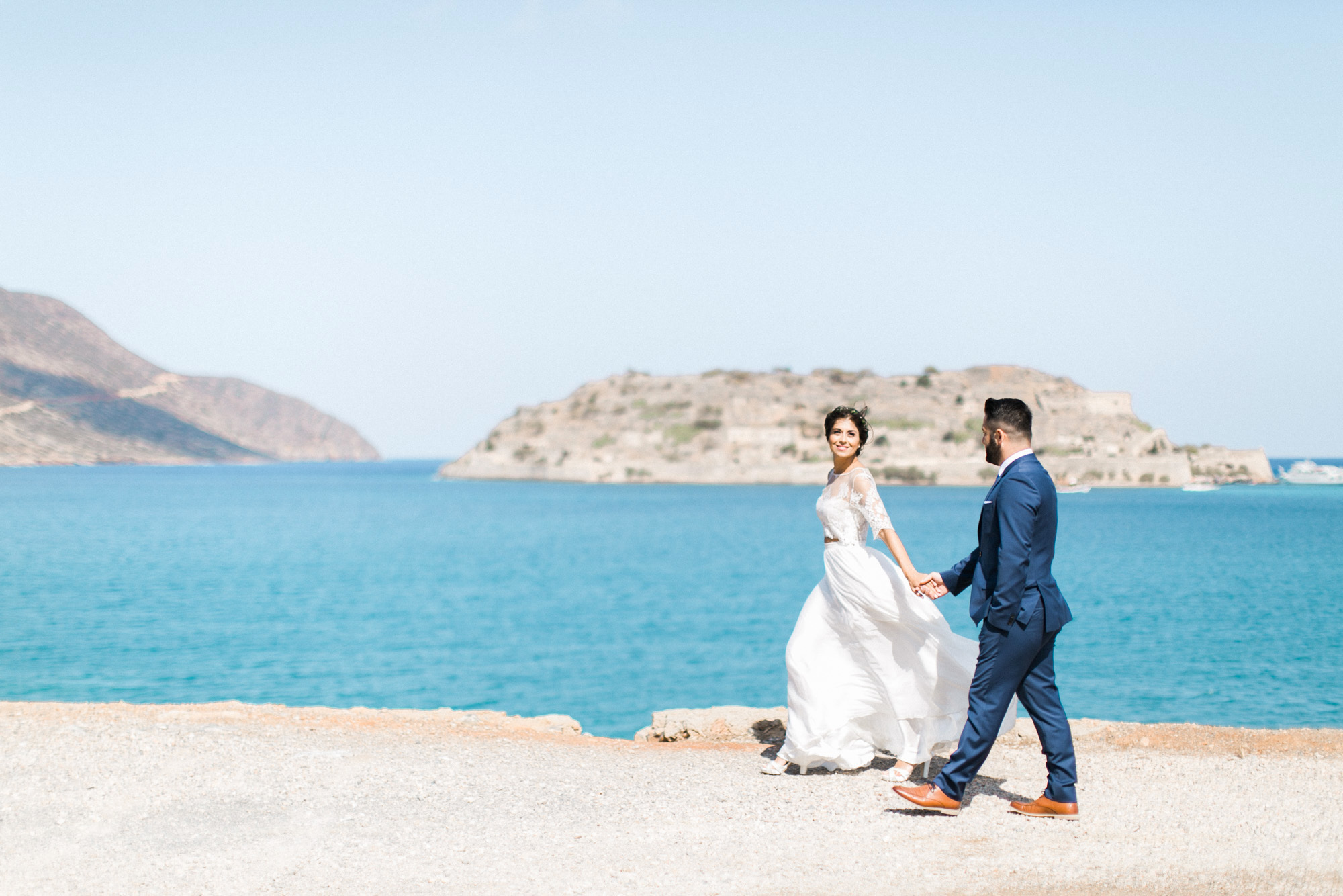 Professional wedding photosession in Elounda Crete, bride and groom walking and posing with Spinalonga island and sea view in the background.
