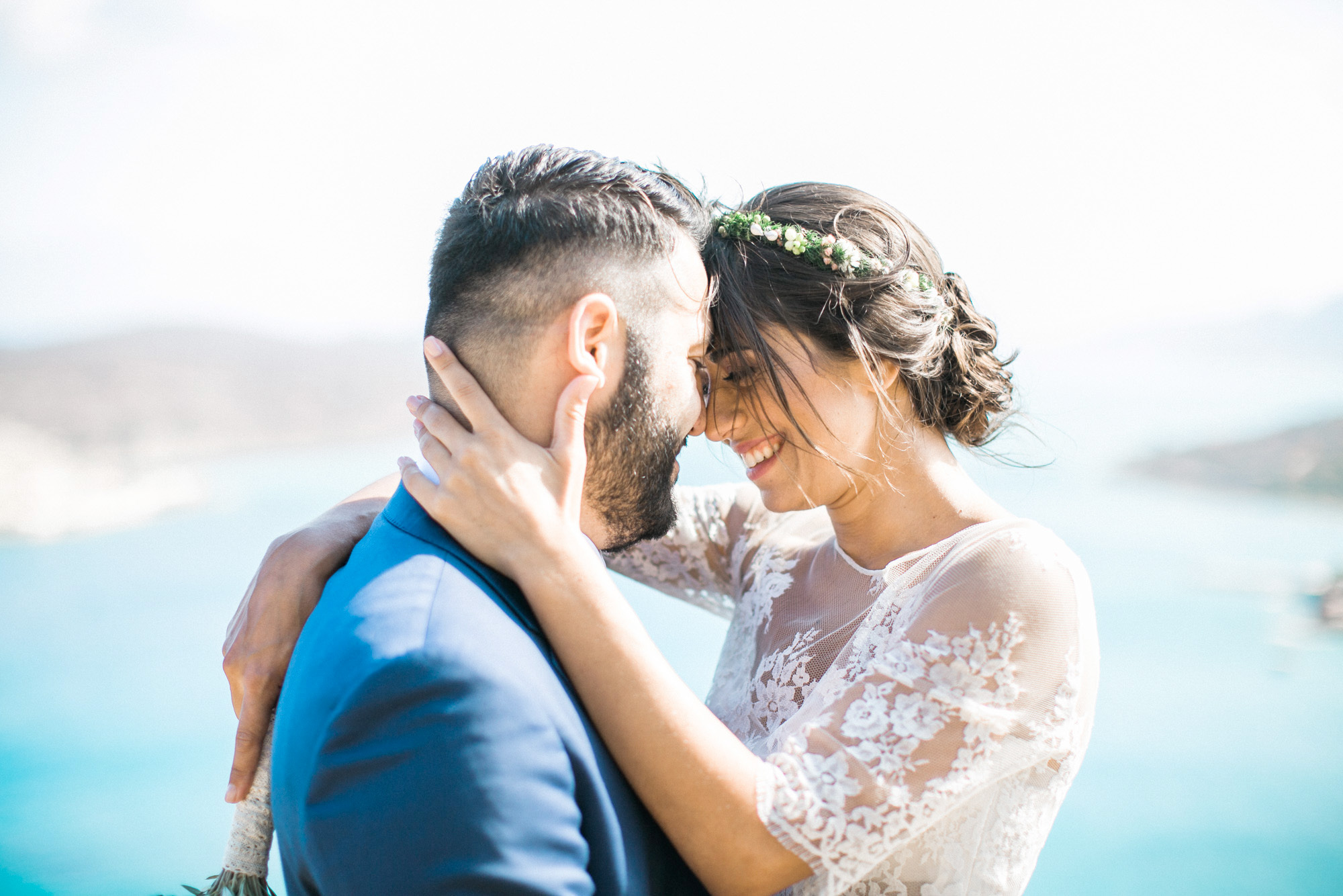 Professional wedding photosession in Crete, closeup portrait of bride and groom posing with Spinalonga island and sea view in the background.