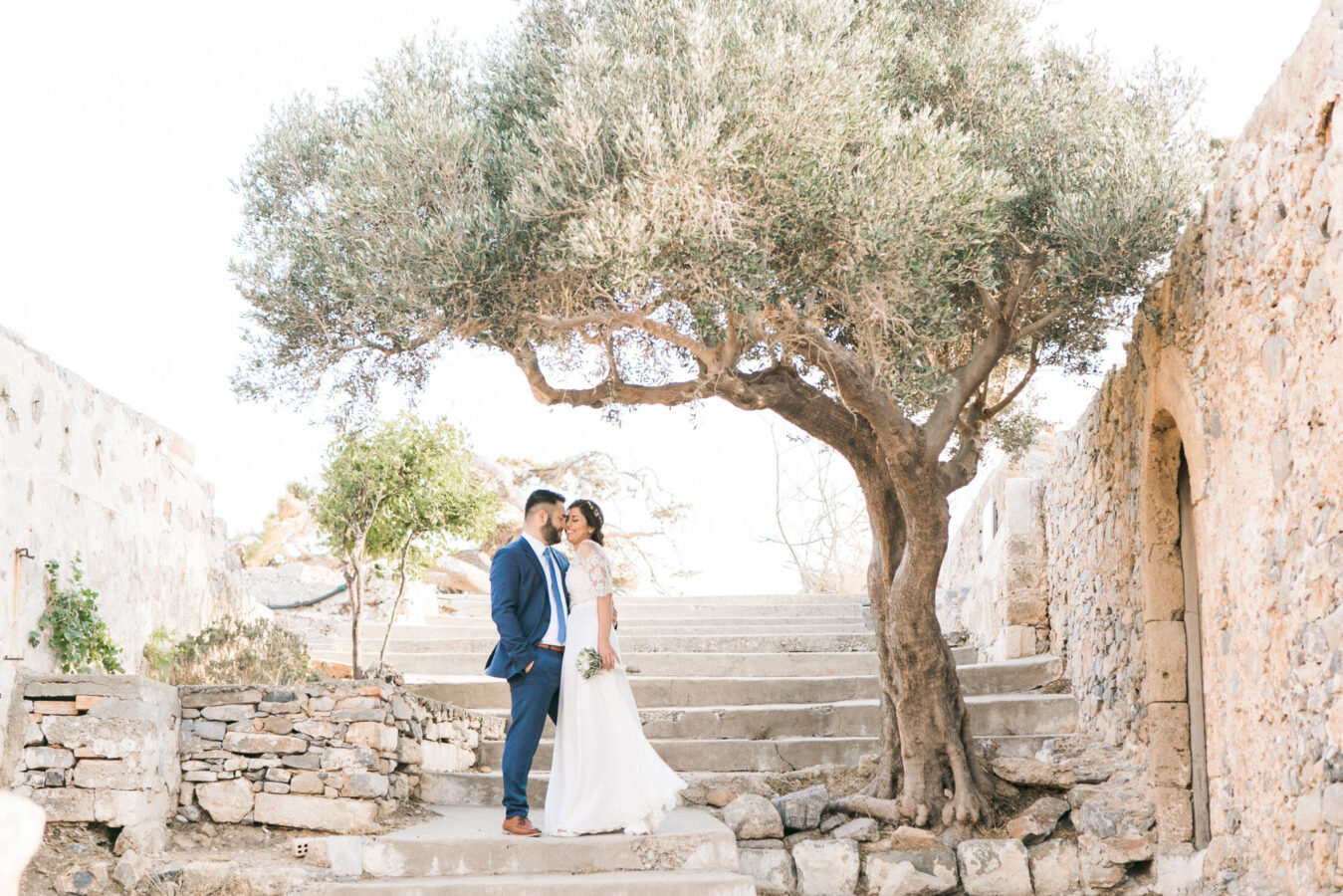 Professional wedding portrait photosession in Crete, bride and groom posing while taking a walk along Spinalonga island, it's historical ruins, church and sites, sea and majestic olive trees in the background.