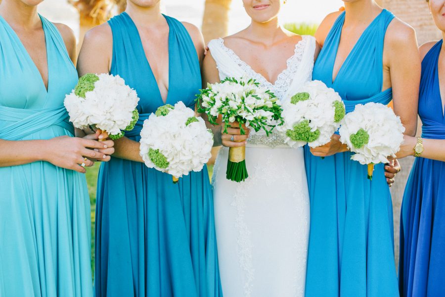 Professional image of a bridal and bridesmaids' bouquets being held by the bride and her friends with the background of white Pronovias dress and blue mismatched dresses by Two Birds bridal boutique.