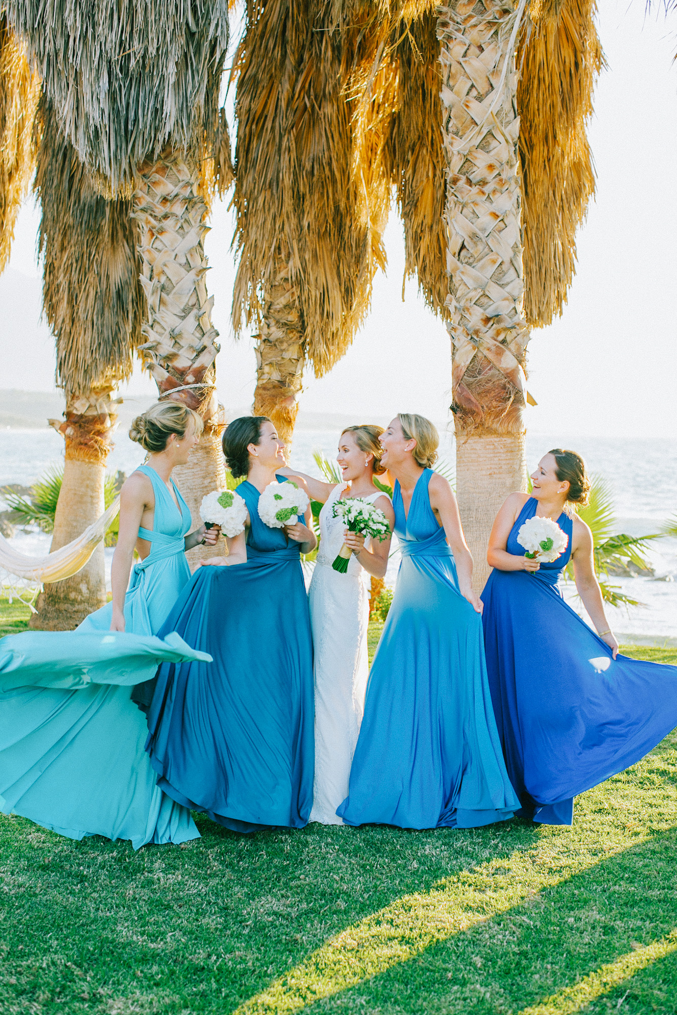 Wedding day portrait of bride and bridesmaids having fun after the wedding ceremony wearing white Pronovias bridal dress and blue mismatched dresses by Two Birds bridal boutique and holding flower bouquets.