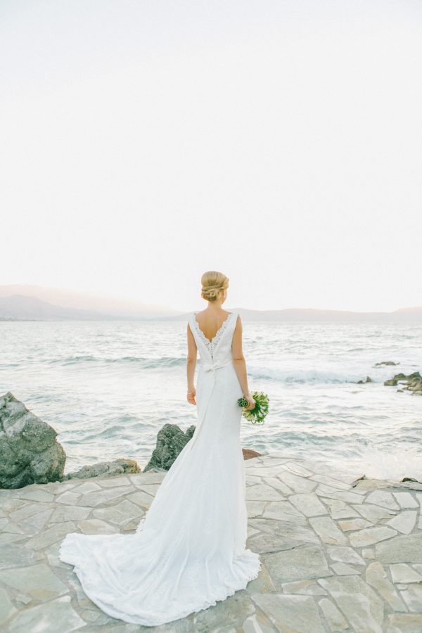 Professional portrait of bride posing for her wedding photographer wearing Pronovias bridal dress and holding flower bouquet captured after the wedding ceremony in palm tree wedding estate in Rethymno Crete with a sunset sea view background.