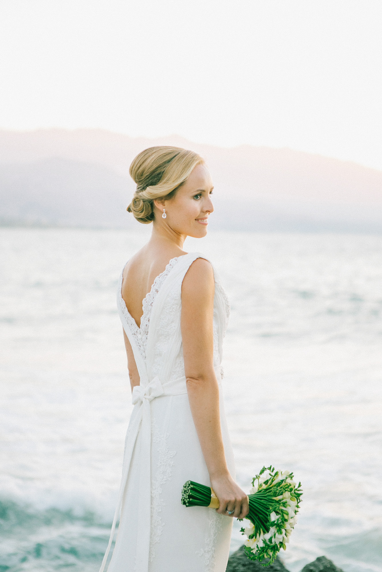 Professional portrait of bride posing for her wedding photographer wearing Pronovias bridal dress and holding flower bouquet captured after the wedding ceremony in palm tree wedding estate in Rethymno Crete with a sunset sea view background.