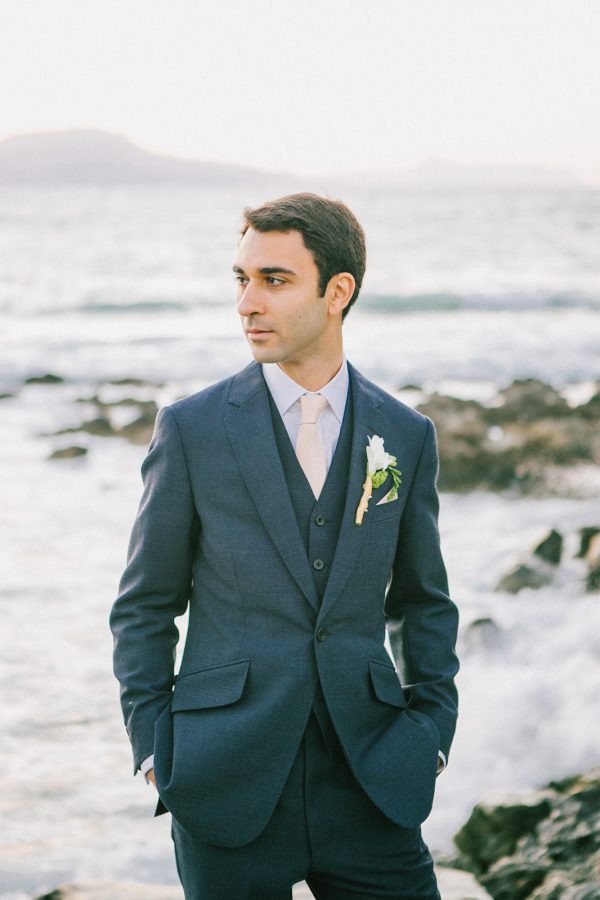 Professional portrait of groom posing for the wedding photographer wearing tailored suit and flower boutonniere captured after the wedding ceremony in palm tree wedding estate in Rethymno Crete with a sunset sea view background.