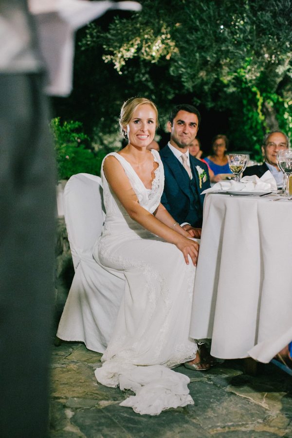 Groom and bride sitting at the head table and listening to speeches during dinner reception at Grecotel Agreco wedding estate in Crete, smiling.
