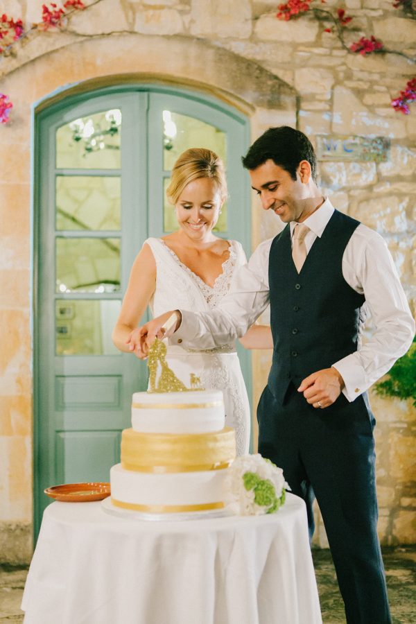 Groom and bride cutting the wedding cake and smiling with the background of stony walls and blue Greek windows of Grecotel Agreco farm.