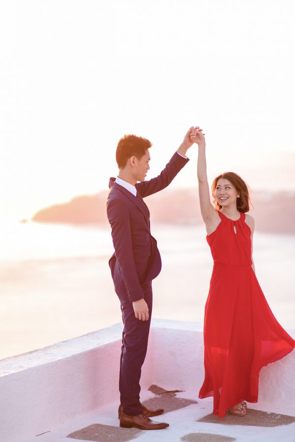 Beautiful Asian couple from Hong Kong is posing for the wedding photographer for their sunset engagement portraits in Imerovigli village Santorini. They're smiling and having good time, wearing formal designer outfits.
