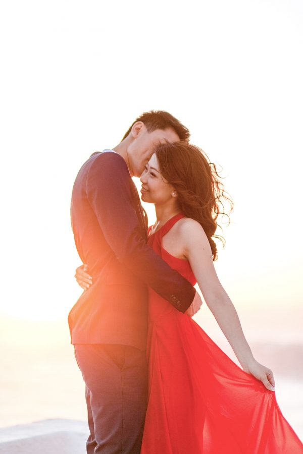 Beautiful Asian couple from Hong Kong is posing for the wedding photographer for their sunset engagement portraits in Imerovigli village Santorini. They're smiling and having good time, wearing formal designer outfits.