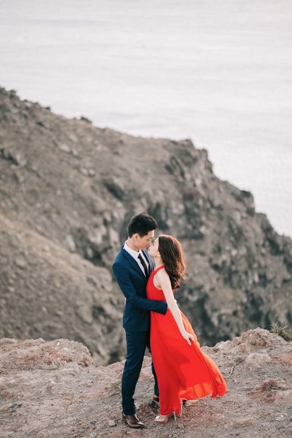 Beautiful Asian couple from Hong Kong is posing for the wedding photographer for their sunset engagement portraits in Imerovigli village Santorini. They're smiling and having good time, wearing formal designer outfits.