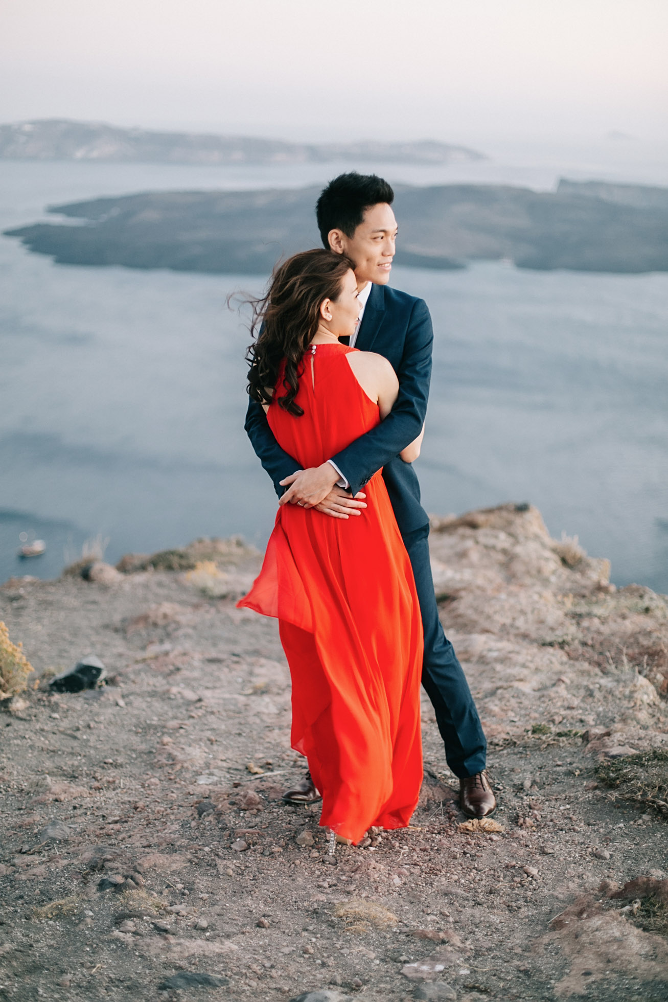 Beautiful Asian couple from Hong Kong is posing for the wedding photographer for their sunset engagement portraits in Imerovigli village Santorini. They're smiling and having good time, wearing formal designer outfits.