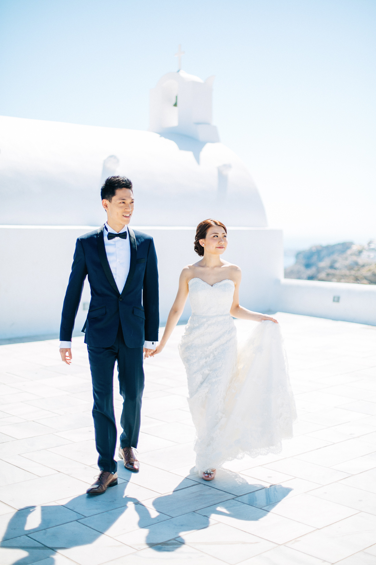 Professional Santorini wedding day photoshoot, groom and bride are walking with the picturesque background of Oia, white church, seaview and clear blue skies.