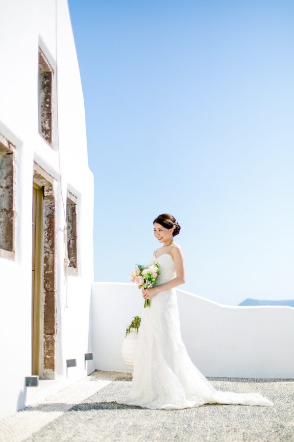 Professional Santorini wedding day photoshoot, bride is posing in front of the church holding flowers with the picturesque background of Oia and clear blue skies.