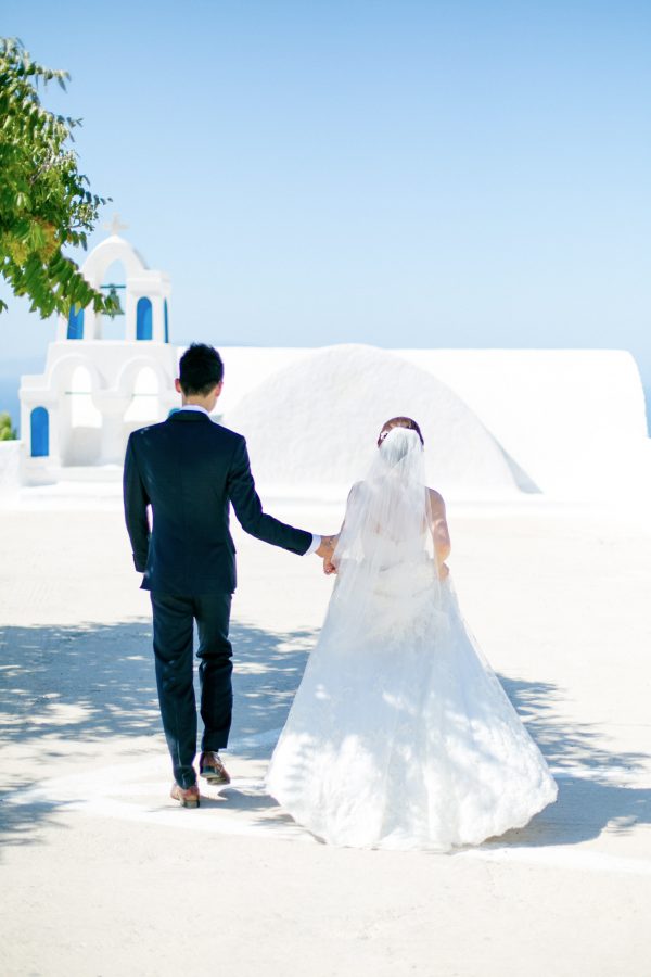 Professional Santorini wedding day photoshoot, groom and bride are walking with the picturesque background of Oia, Santorini church and clear blue skies.