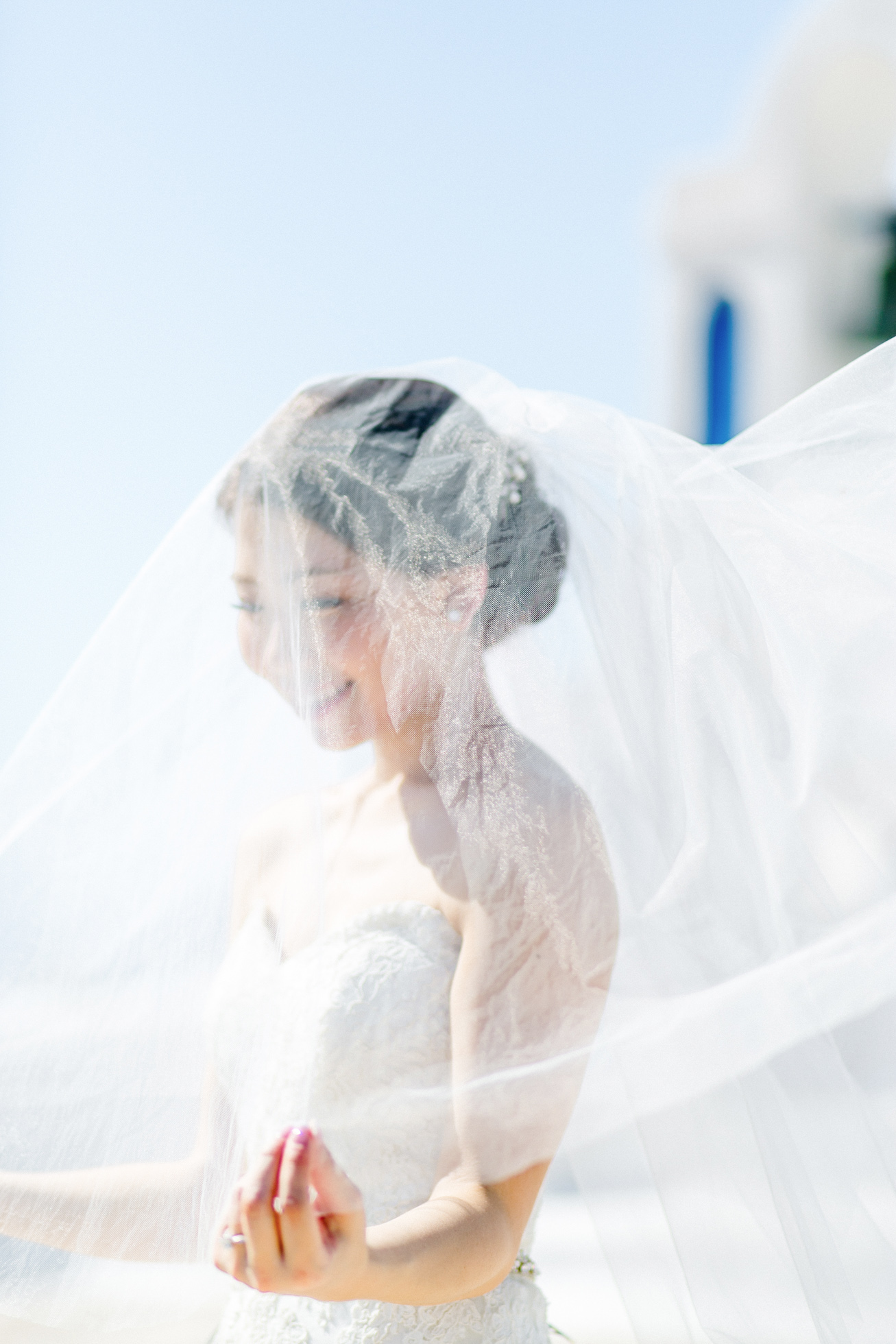 Professional Santorini wedding day photoshoot, bride is posing in front of the church holding her flowing veil with the picturesque background of the wedding chapel and clear blue skies.
