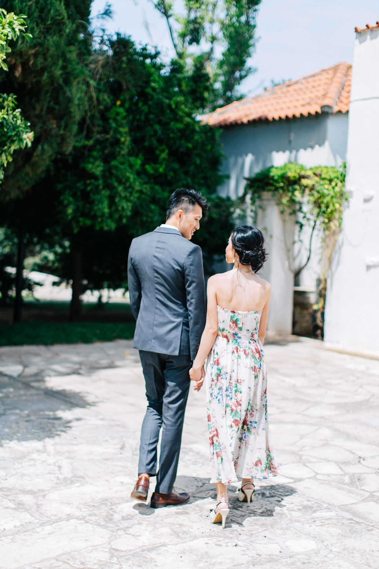 Young elegant couple wearing summer outfits is posing for their elopemement portraits for professional photographer team in Crete island, Greece. Colorful garden and park backdrop adds to the happy and vibrant feel of this session.