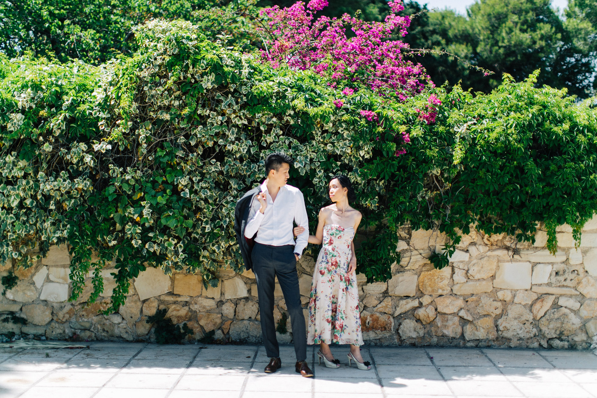 Young elegant couple wearing summer outfits is posing for their elopemement portraits for professional photographer team in Crete island, Greece. Colorful garden and park backdrop adds to the happy and vibrant feel of this session.