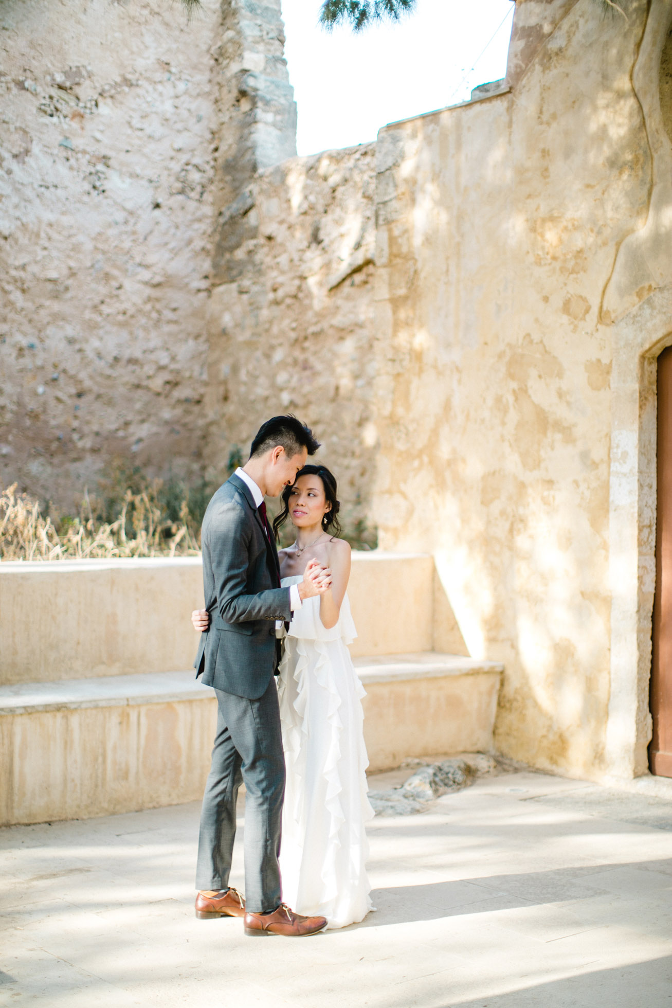 Elegant Asian couple wearing designer wedding clothing posing for professional photographer team during their wedding day photoshoot in Fortezza of Rethymno town, Crete.