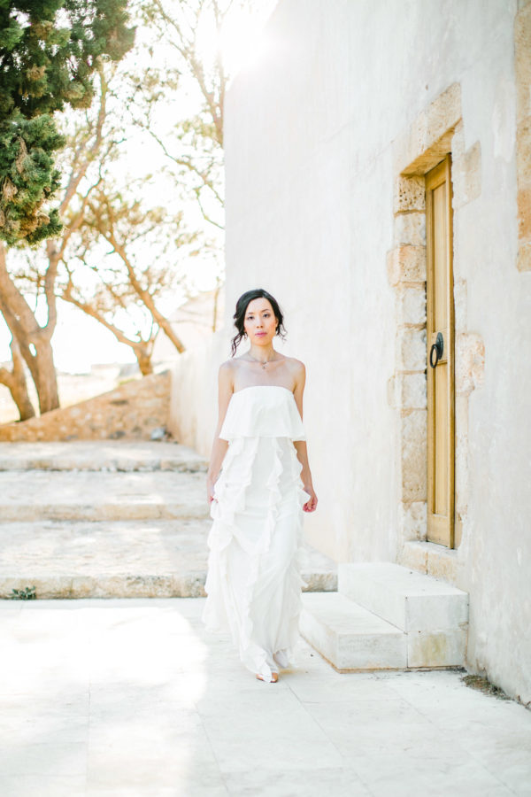 Elegant Asian bride wearing white understated and posh bridal dress posing for professional photographer for her wedding portraits in Fortezza of Rethymno town, Crete.