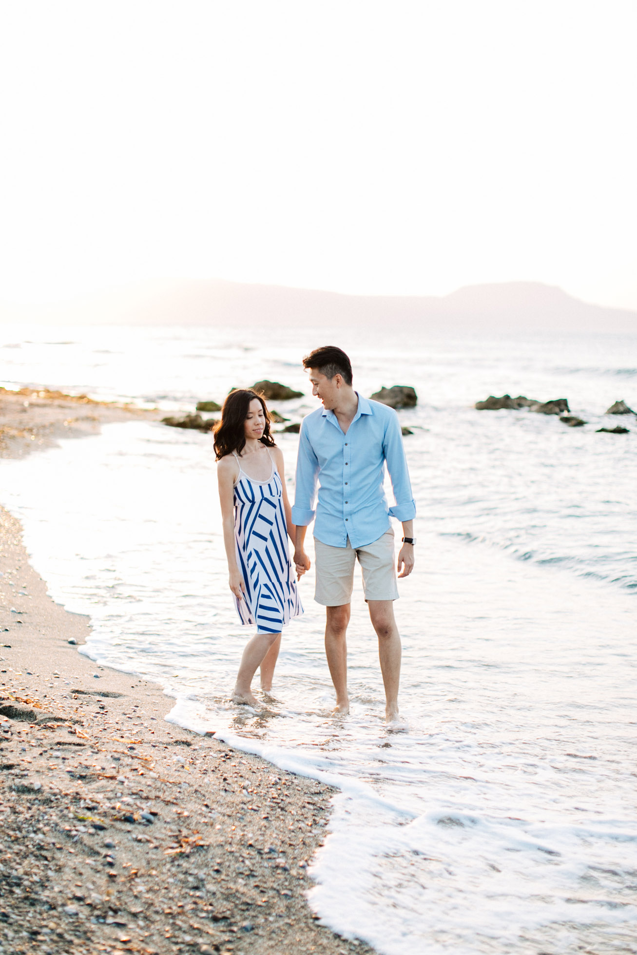 Happy international couple wearing summer clothing is posing for professional photographer team during their beach wedding photoshoot on the secluded shores of Rethymno town, Crete, Greece.