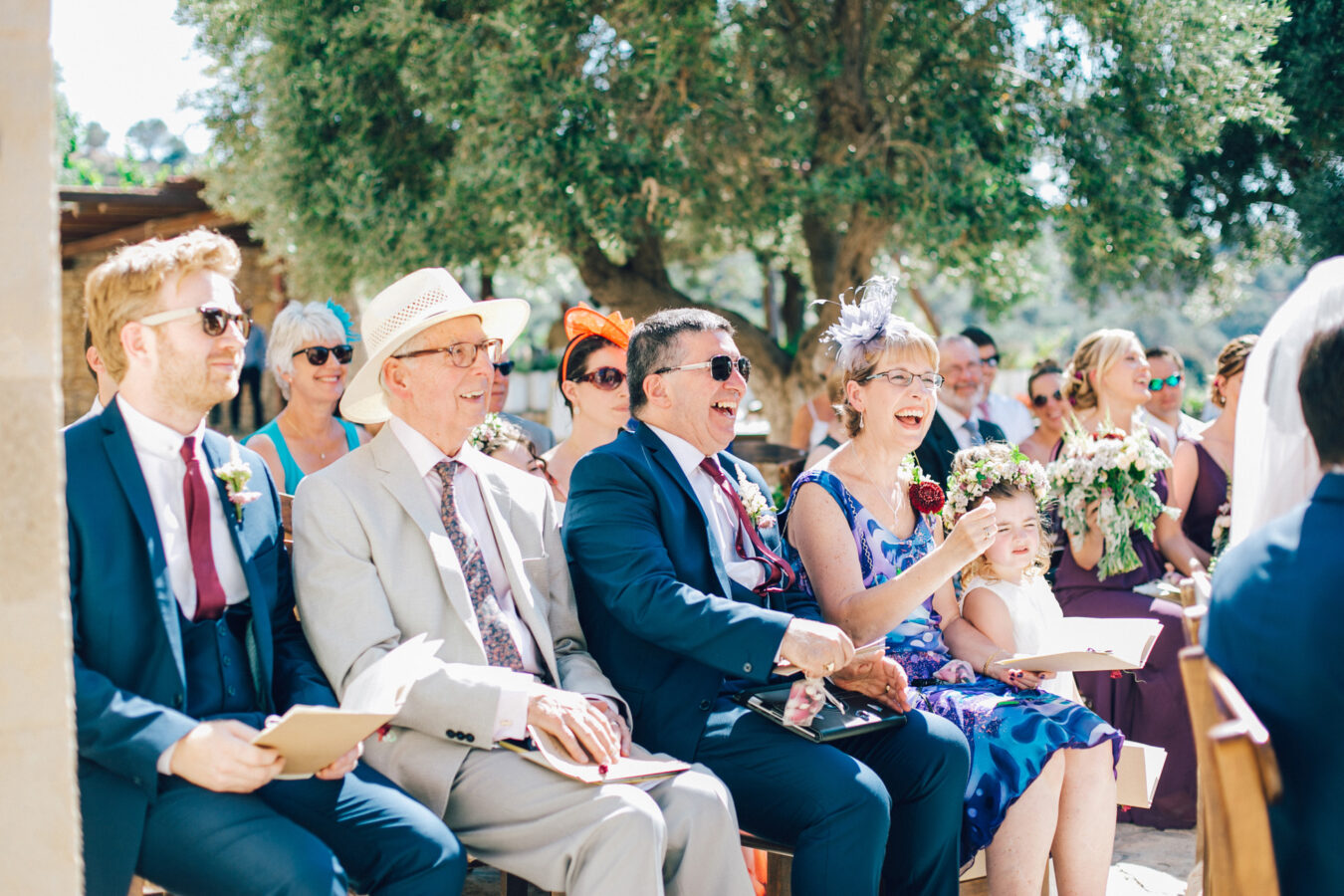 Candid image of a happy reaction of the wedding guests crowd on a destination wedding day in Agreco Farm in Crete, Greece, captured by wedding photographer. Guests are smiling and laughing.